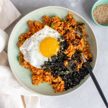 A bowl of tuna kimchi fried rice with a fried egg and shredded seaweed with a spoon.