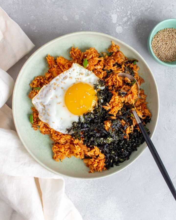 A bowl of tuna kimchi fried rice with a fried egg and shredded seaweed with a spoon.
