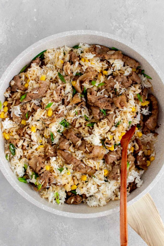 A pan with stir fried sliced beef, rice, and corn.