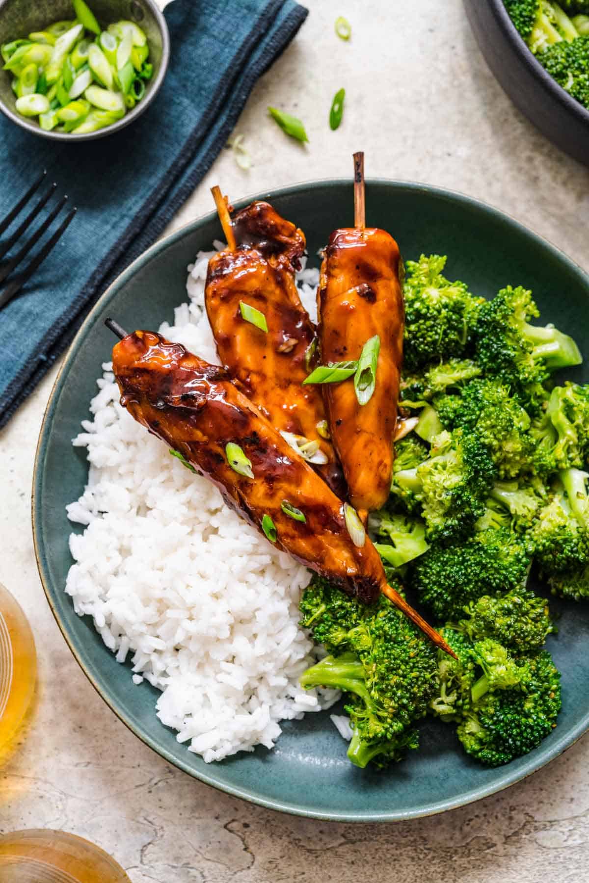 A blue plate with rice and broccoli with three teriyaki chicken skewers on top.