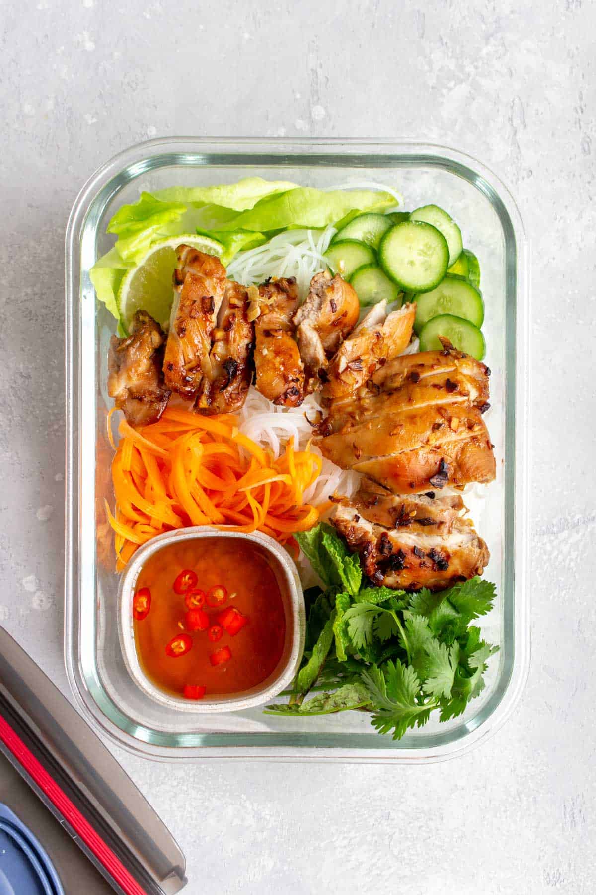 Overhead view of a meal prep container of air fryer lemongrass chicken, noodles, carrots, cilantro, cucumber, and nuoc cham.