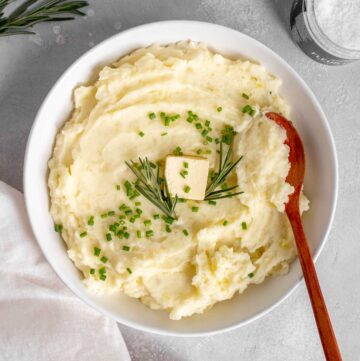 A bowl of rosemary mashed potatoes with a knob of butter, chives, and rosemary on top.
