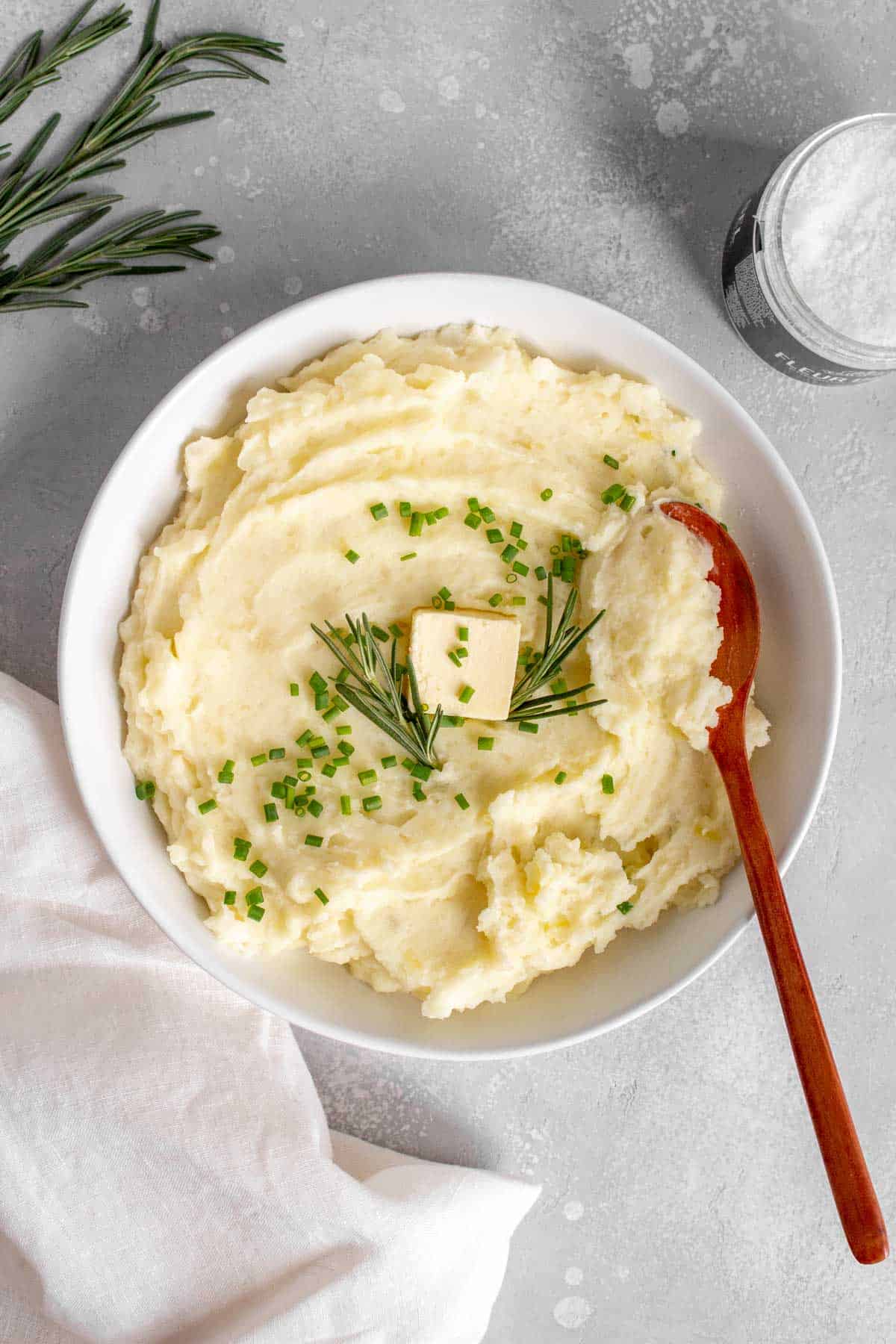 A bowl of rosemary mashed potatoes with a knob of butter, chives, and rosemary on top.