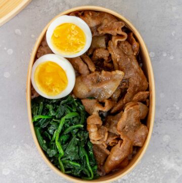 Overhead view of a bento box with cooked sliced pork, spinach, and eggs over rice.