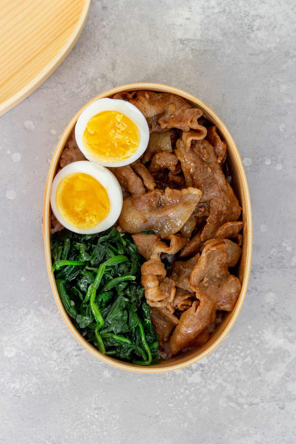 Overhead view of a bento box with cooked sliced pork, spinach, and eggs over rice.