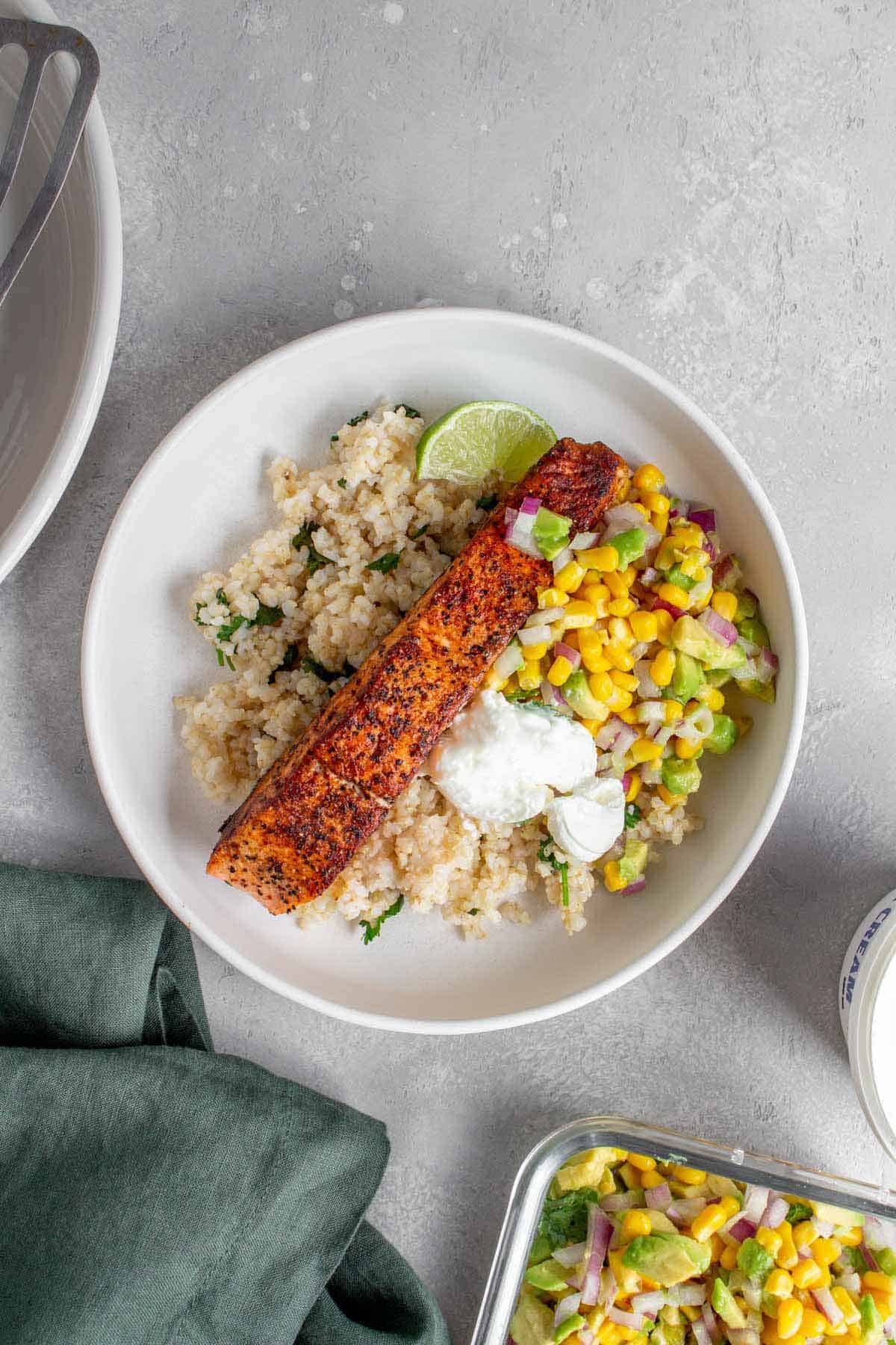 Overhead view of a plate of cilantro lime brown rice, avocado corn salsa, sour cream, lime wedge, and pan seared salmon.