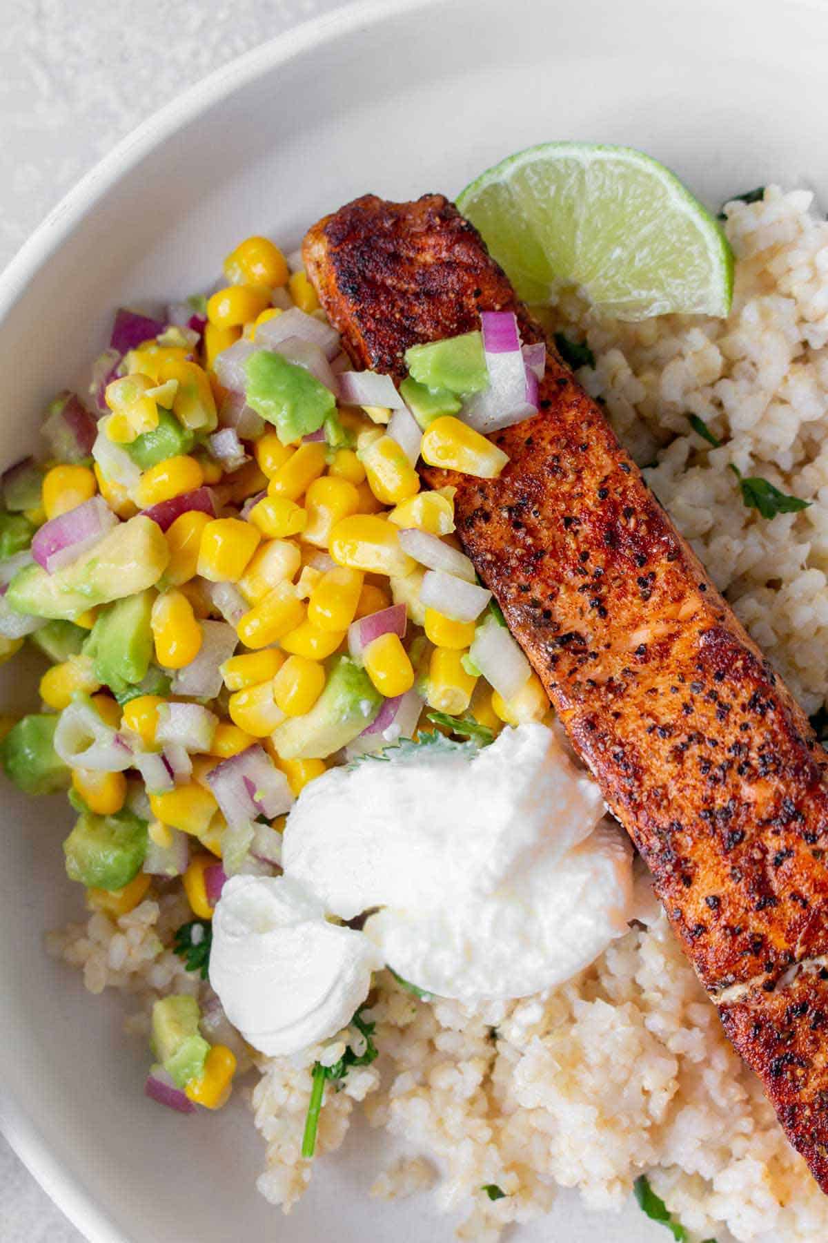 Close up of pan seared salmon over rice with avocado corn salsa and sour cream.
