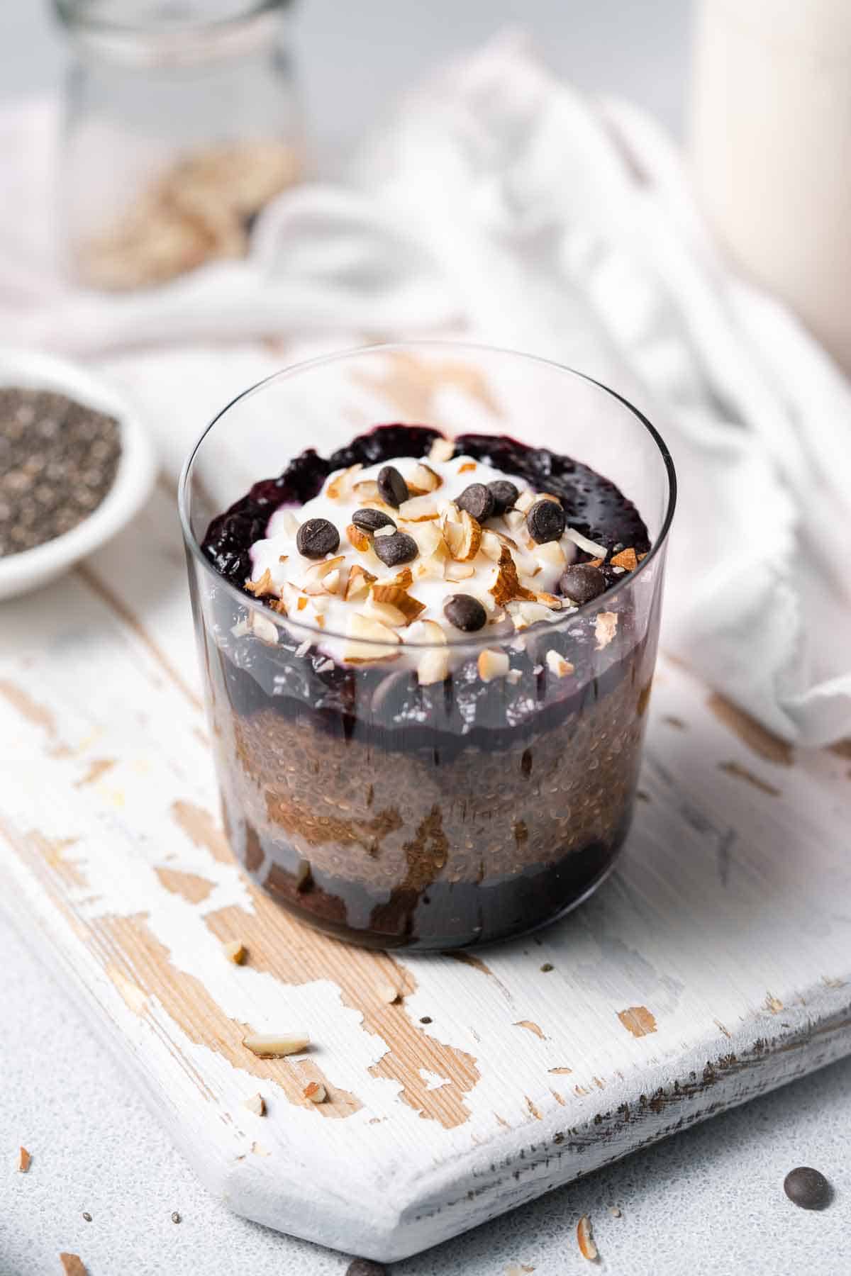 An angled view of a glass of chocolate blueberry chia pudding topped with yogurt and chocolate chips.