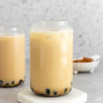 A glass of jasmine milk tea on a coaster in front of a second glass in the background.