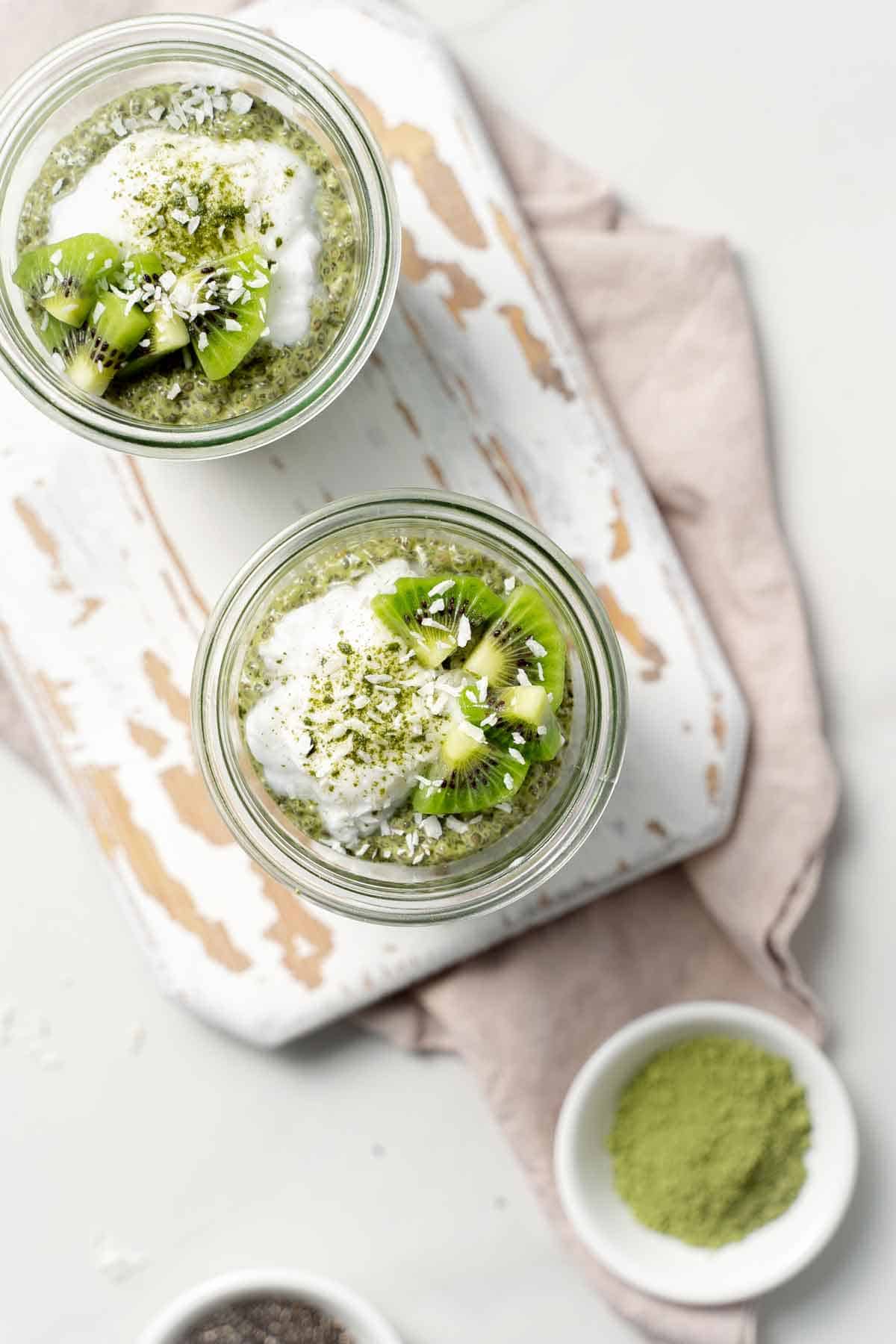 Overhead view of two glasses of matcha chia pudding topped with yogurt, kiwi, and coconut.