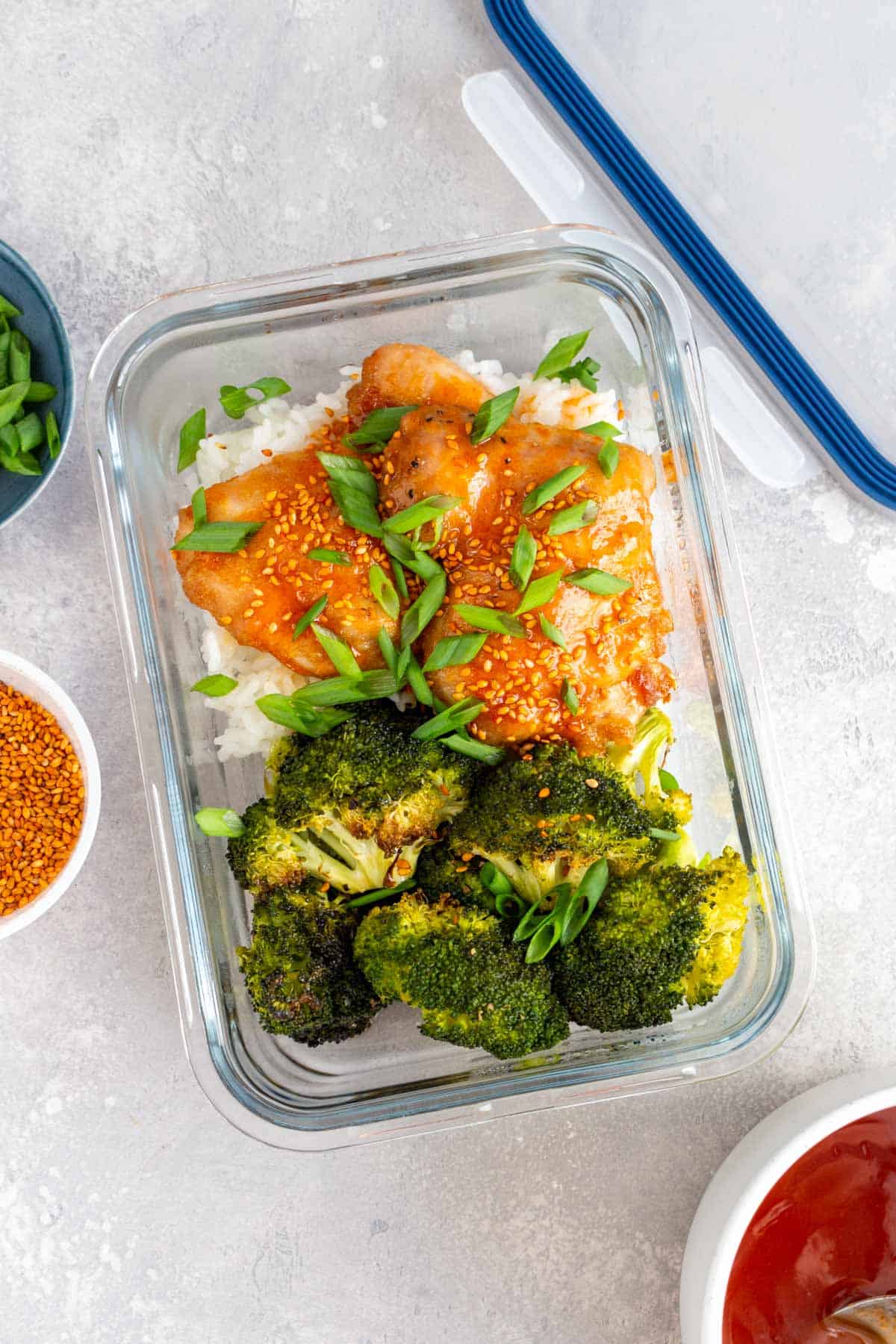 A meal prep container with sheet pan chicken thighs and broccoli with honey sriracha glaze over rice garnished with green onions.