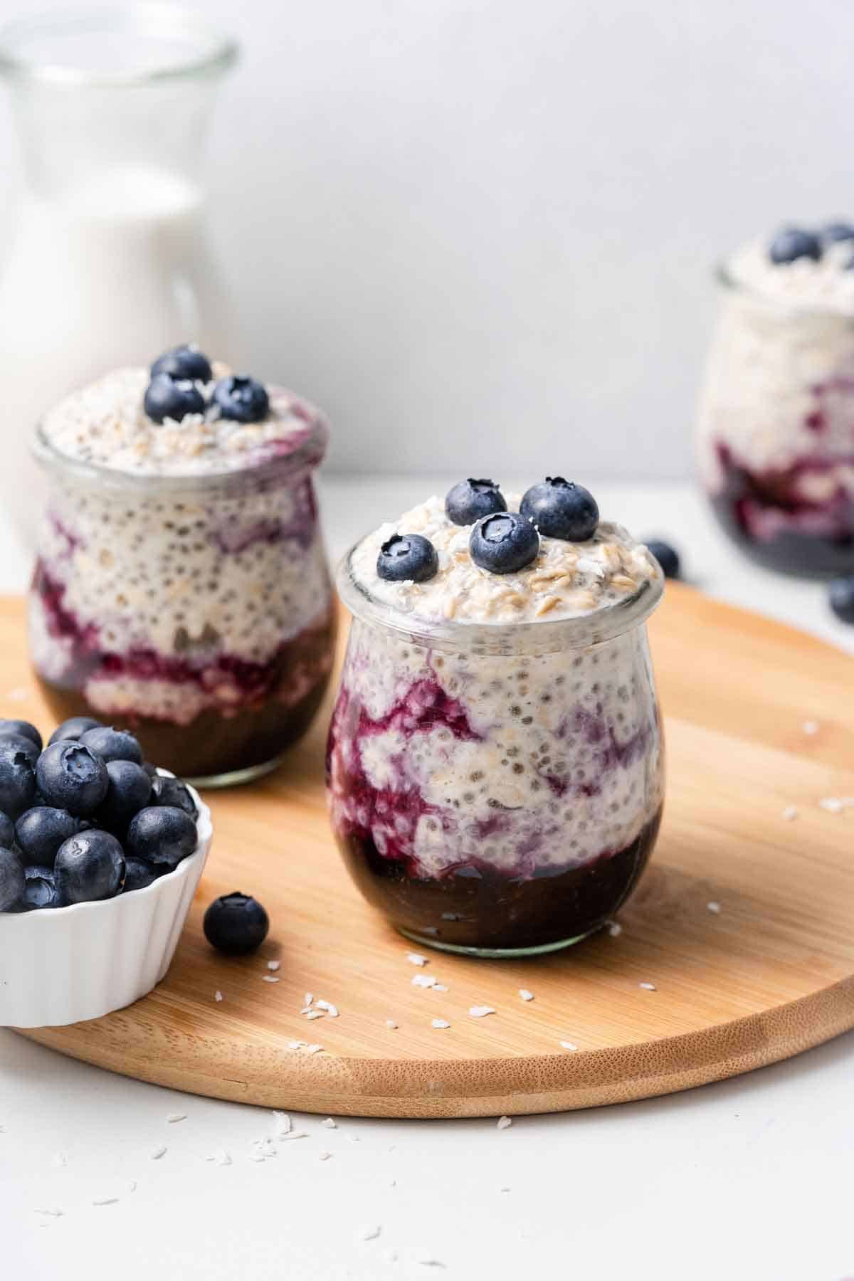 A jar of blueberry overnight oats with two in the background with a small bowl of blueberries on the side.