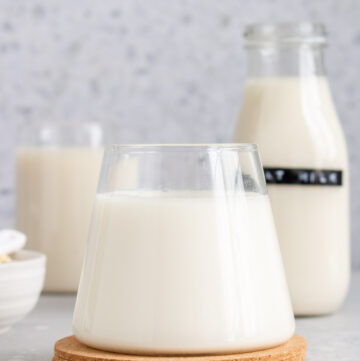 Profile view of a glass of oat milk with a bottle in the background.