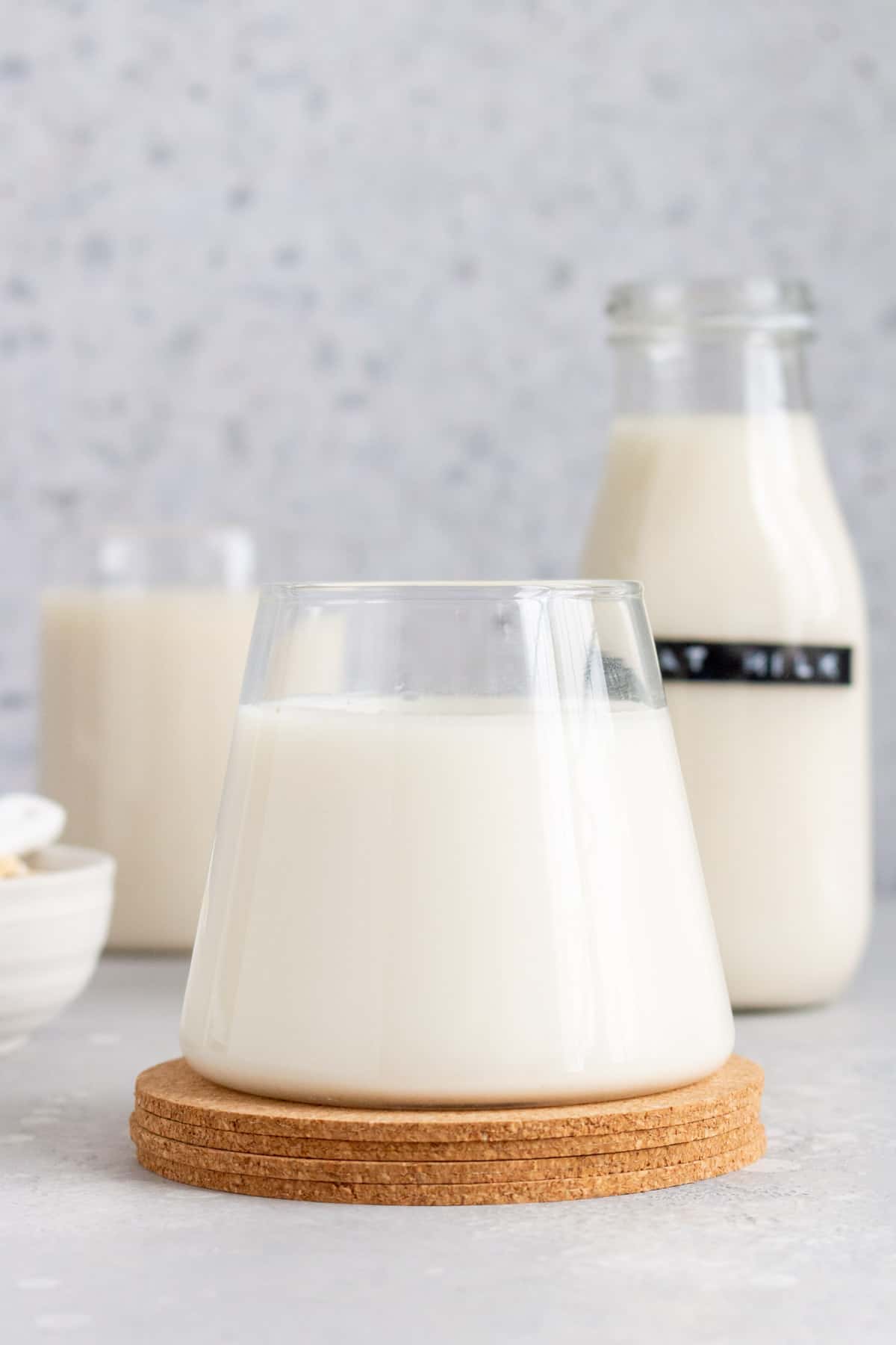 Profile view of a glass of oat milk with a bottle in the background.