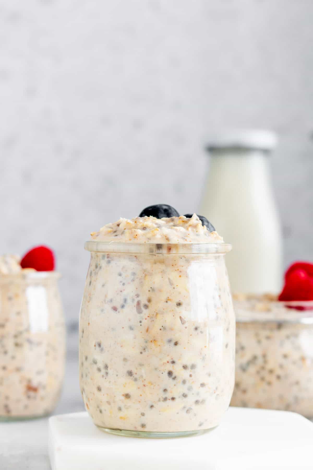 Close view of a jar of overnight oats with almond milk with blueberries on top with more jars in the back with raspberries on top.