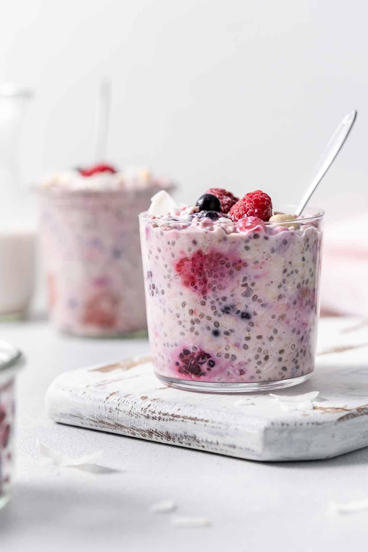 A glass of overnight oats with frozen fruit with a spoon inside with another jar out of focus in the back.