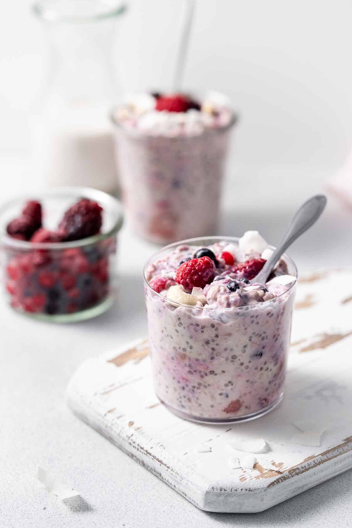 A jar of overnight oats with frozen fruit with a spoon inside with a small jar of mixed berries and another jar in the background.