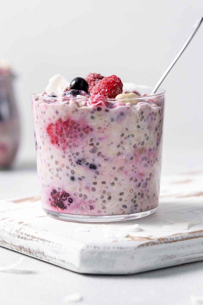 A jar of overnight oats with frozen fruit with a spoon inside and mixed berries on top.