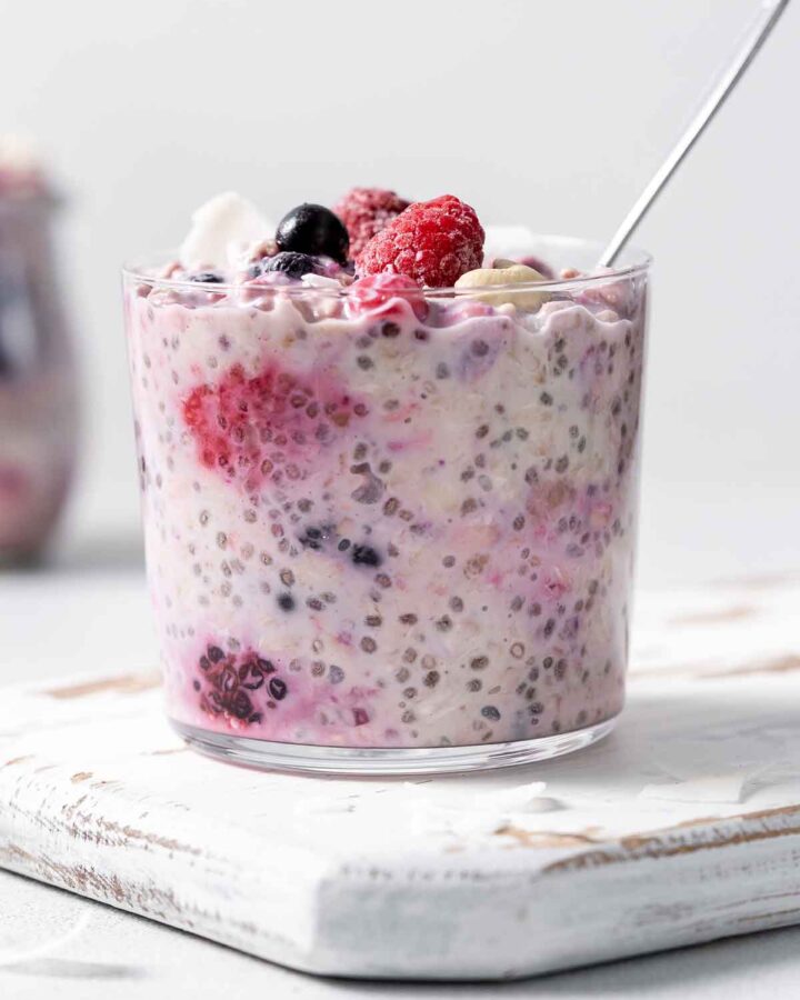 A jar of overnight oats with frozen fruit with a spoon inside and mixed berries on top.