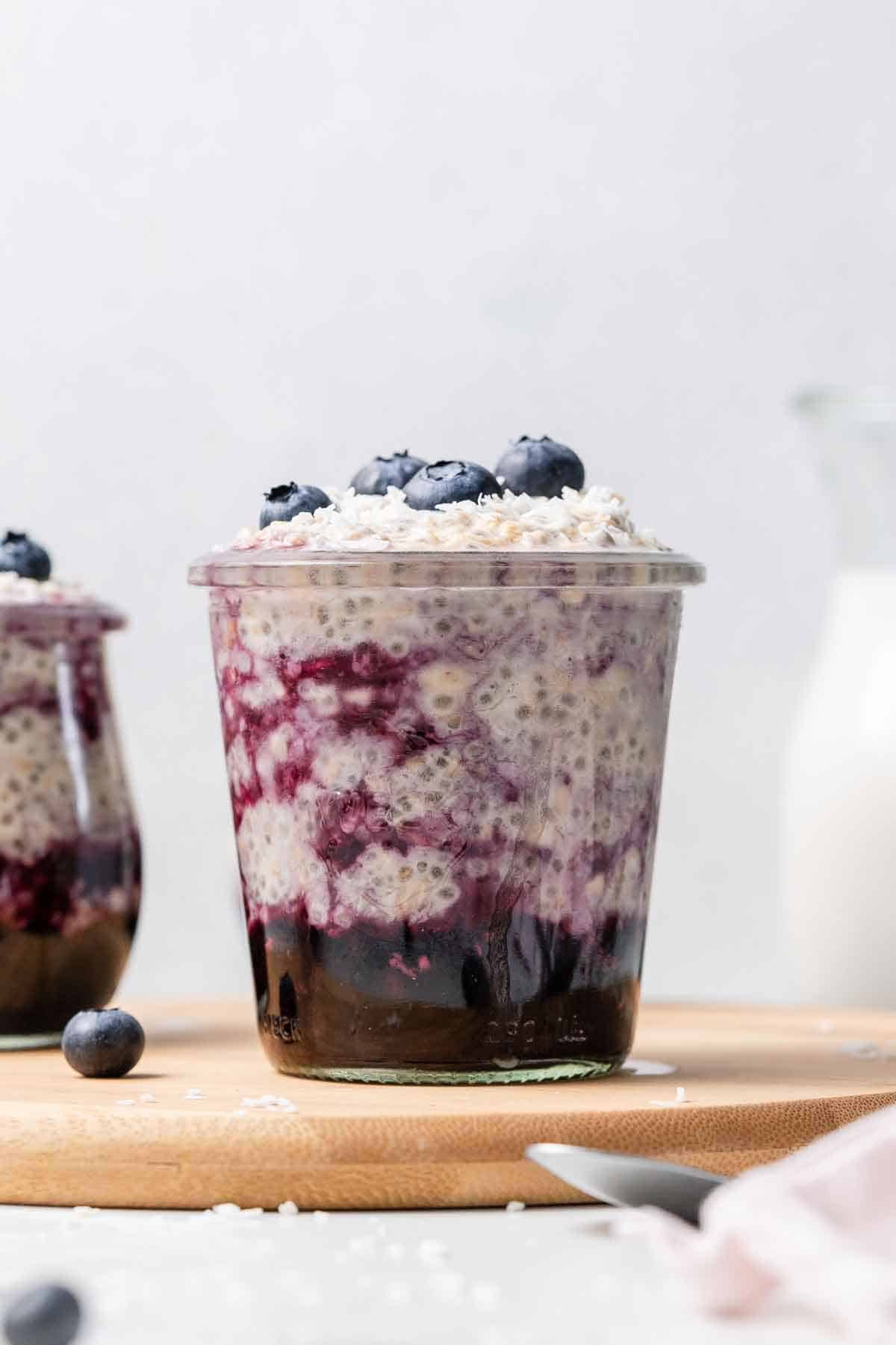 Profile view of a glass of overnight oats with water with some cooked blueberries mixed into the bottom and topped with fresh blueberries.