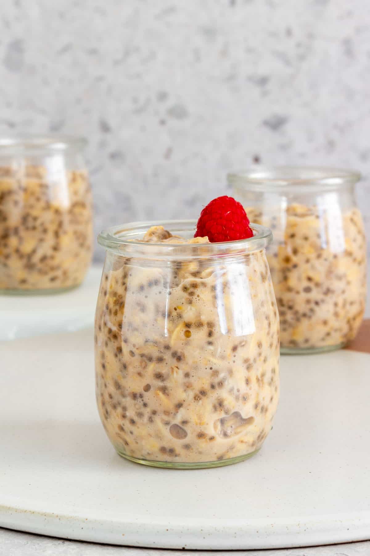 A cup of applesauce overnight oats with a fresh raspberry on top with two more cups in the background.