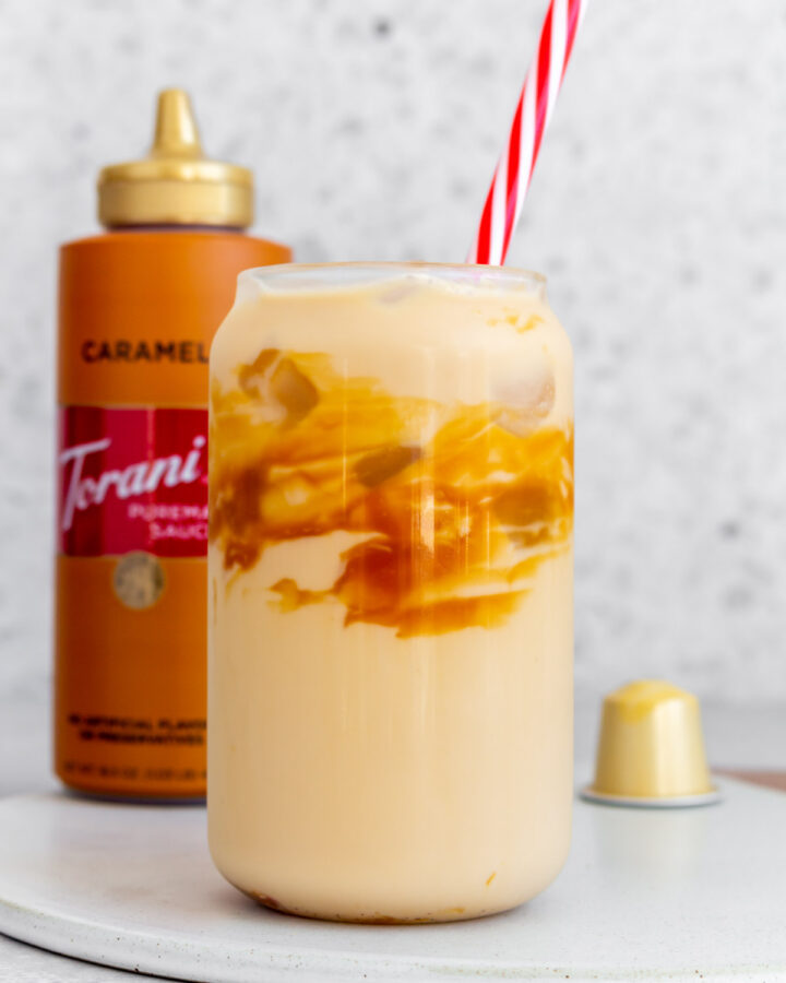 A glass of caramel macchiato with caramel sauce on the glass with a straw. A bottle of caramel sauce in the background.