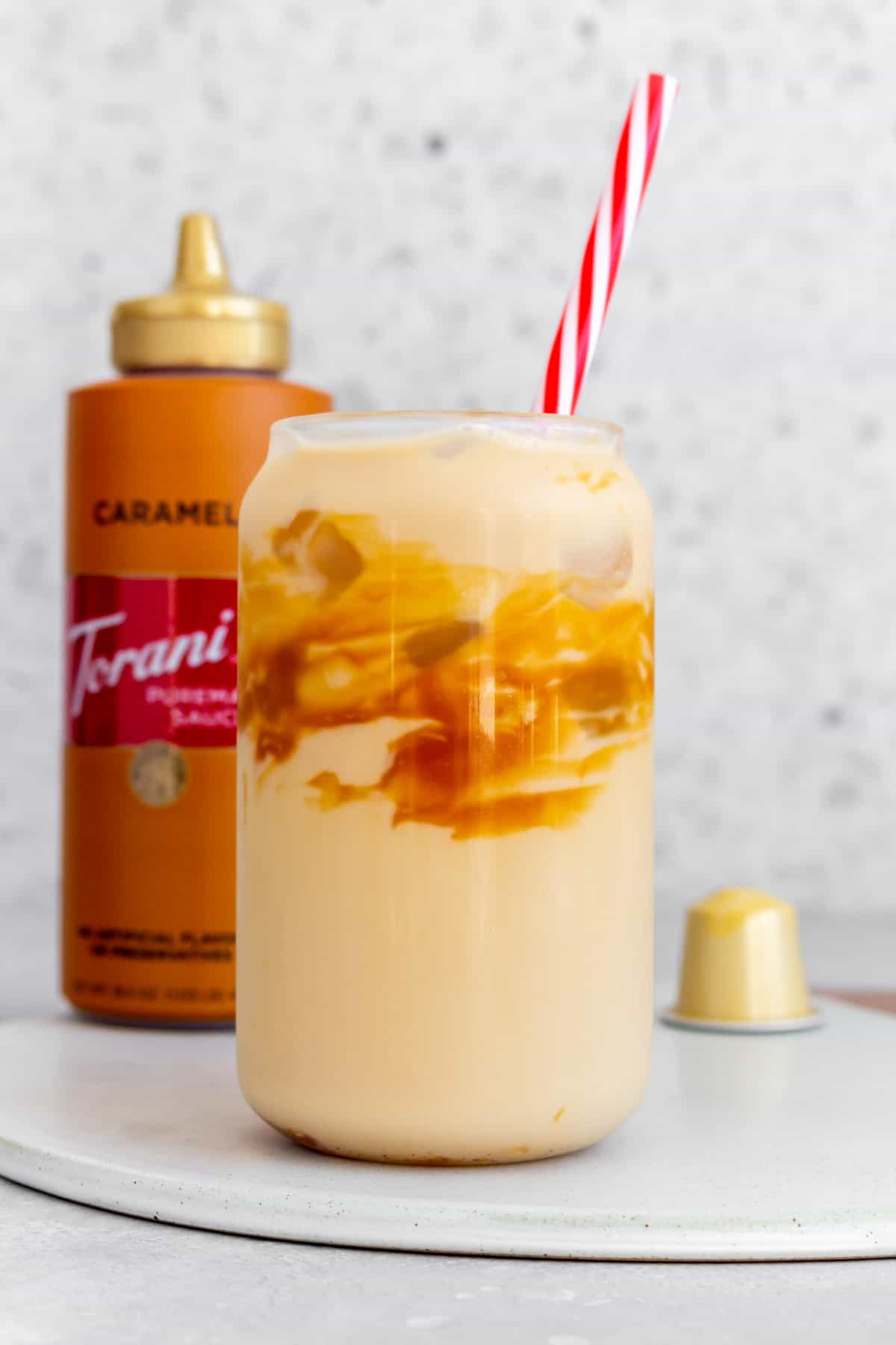 A glass of caramel macchiato with caramel sauce on the glass with a straw. A bottle of caramel sauce in the background.