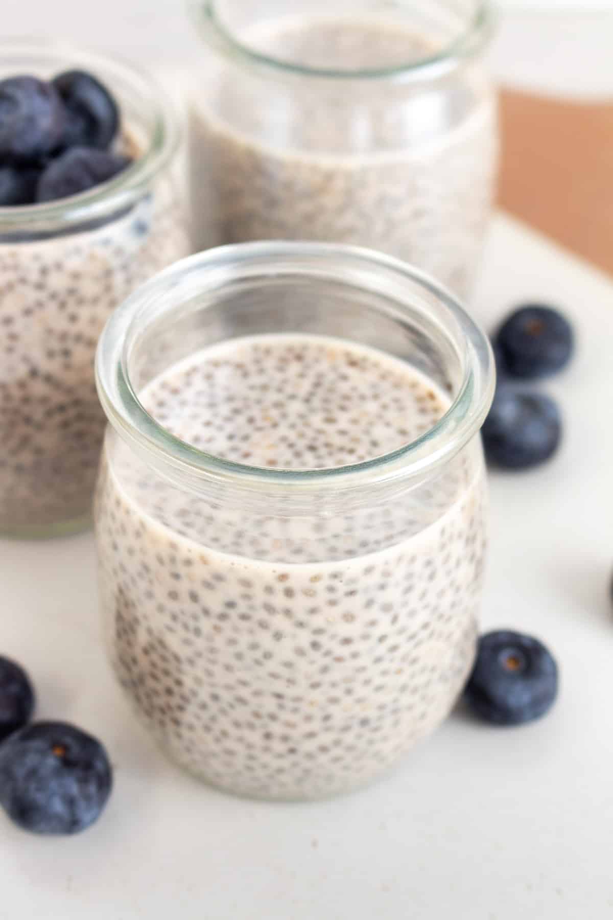A cup of protein chia pudding with two more in the background and blueberries scattered around.