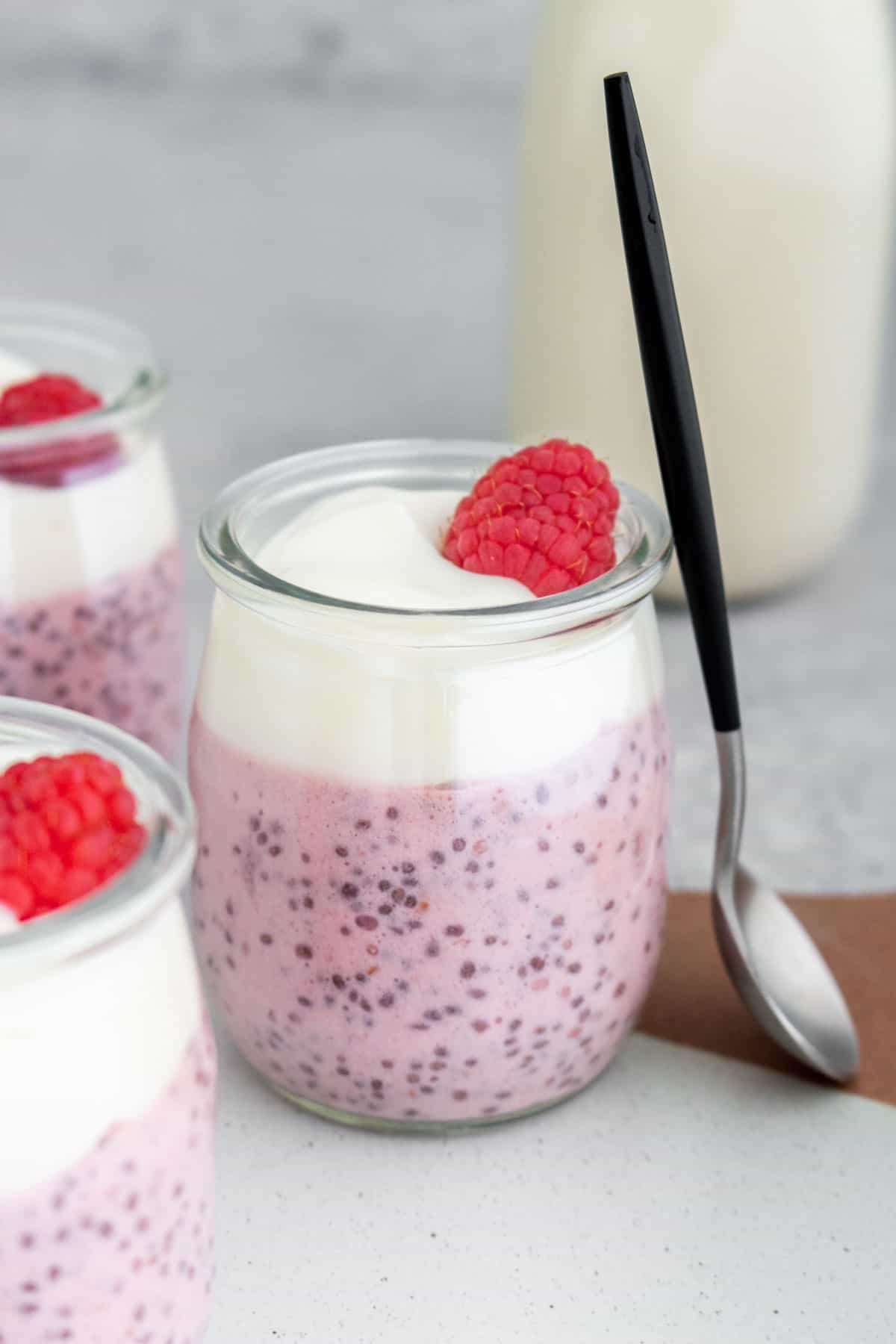 A cup of raspberry chia pudding topped with yogurt and a raspberry with a spoon leaning up against it.