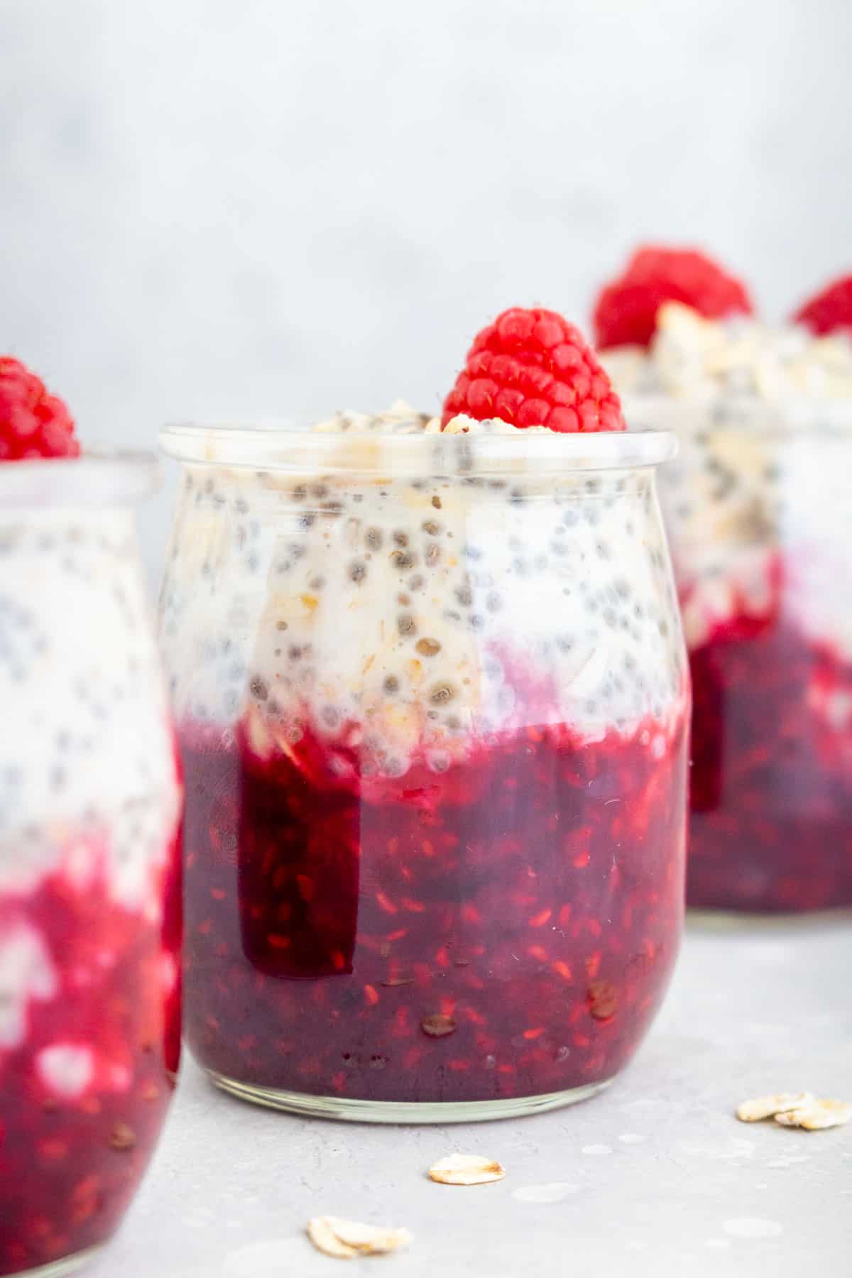 A jar of raspberry overnight oats with a fresh raspberry on top sandwiched between two jars.