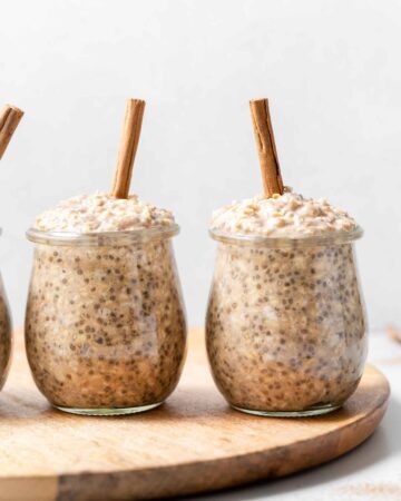 Two jars of cinnamon overnight oats with cinnamon sticks on top on a wooden serving board.