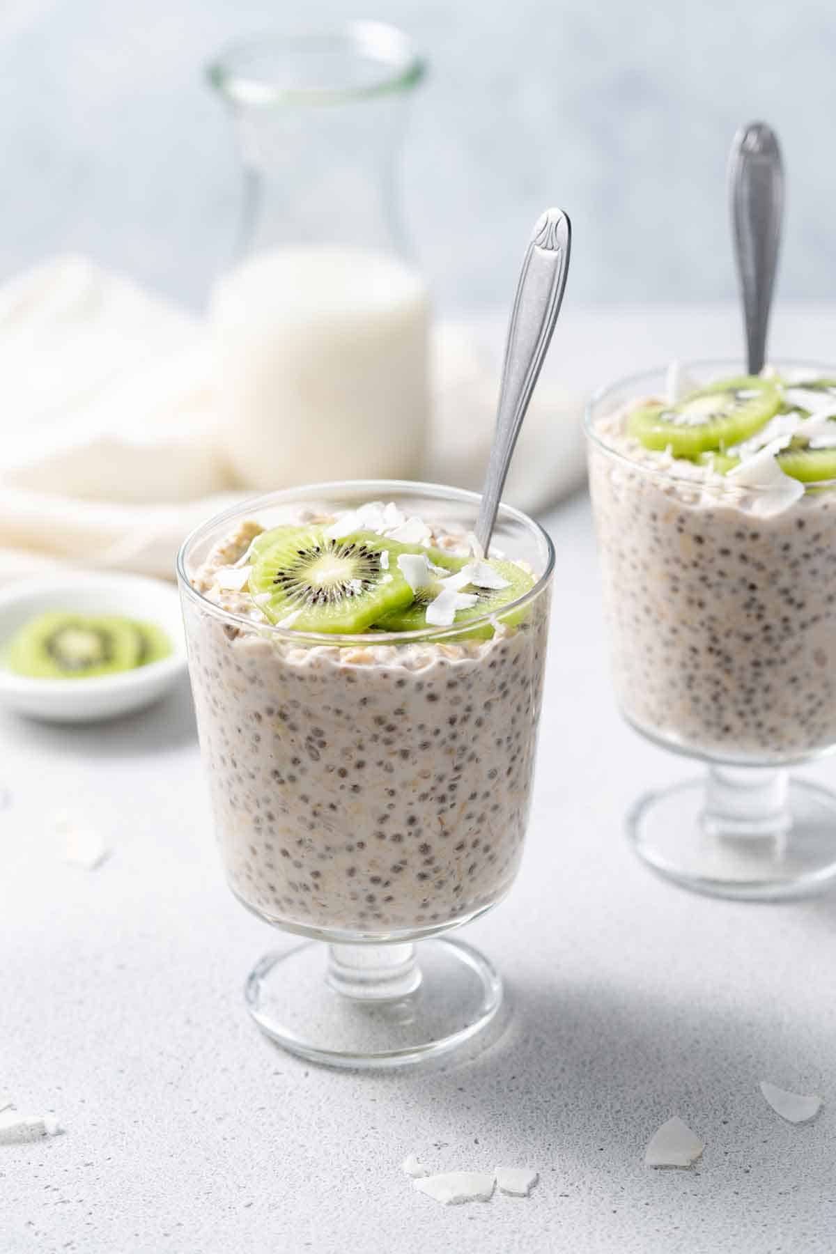 A glass of coconut overnight oats with kiwi and coconut on top with a spoon inside.