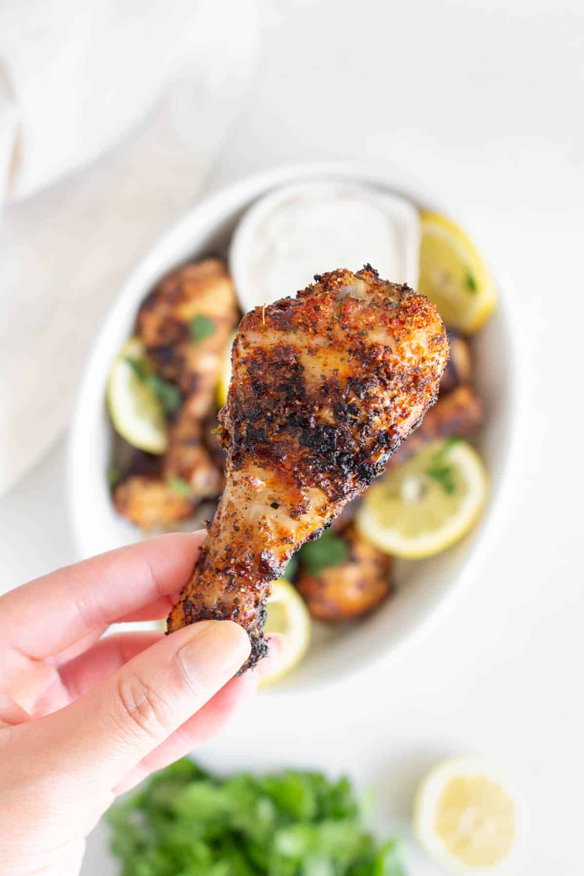 A hand holding up a grilled chicken drumstick over a platter of more drumsticks.
