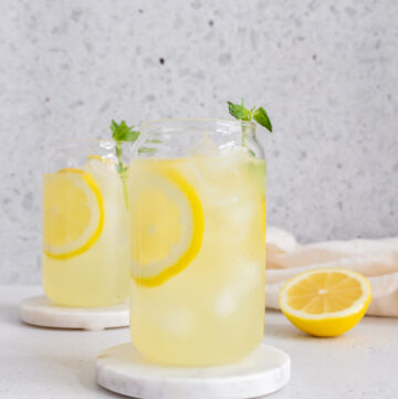 A glass of basil lemonade on a marble coaster with a second glass in the background along with a cut lemon.