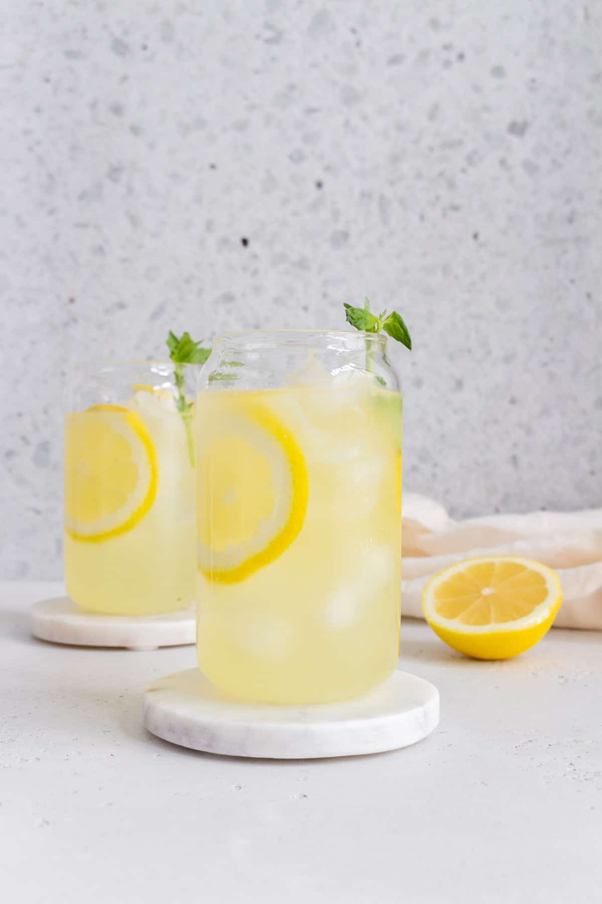 A glass of basil lemonade on a marble coaster with a second glass in the background along with a cut lemon.