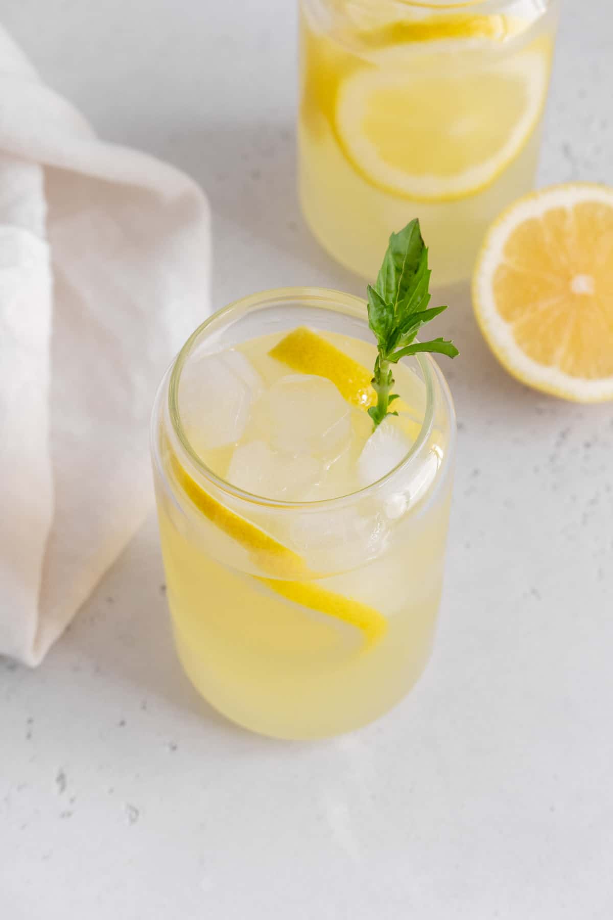 A glass of basil lemonade with basil garnish and a second glass with a cut lemon in the background.