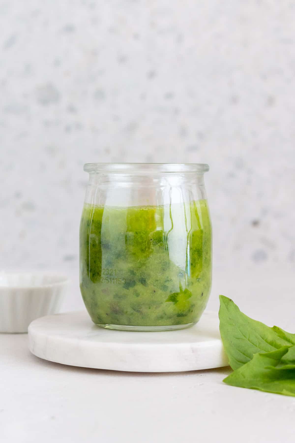 Profile view of a jar of basil vinaigrette on a marble coaster with some basil on the side.