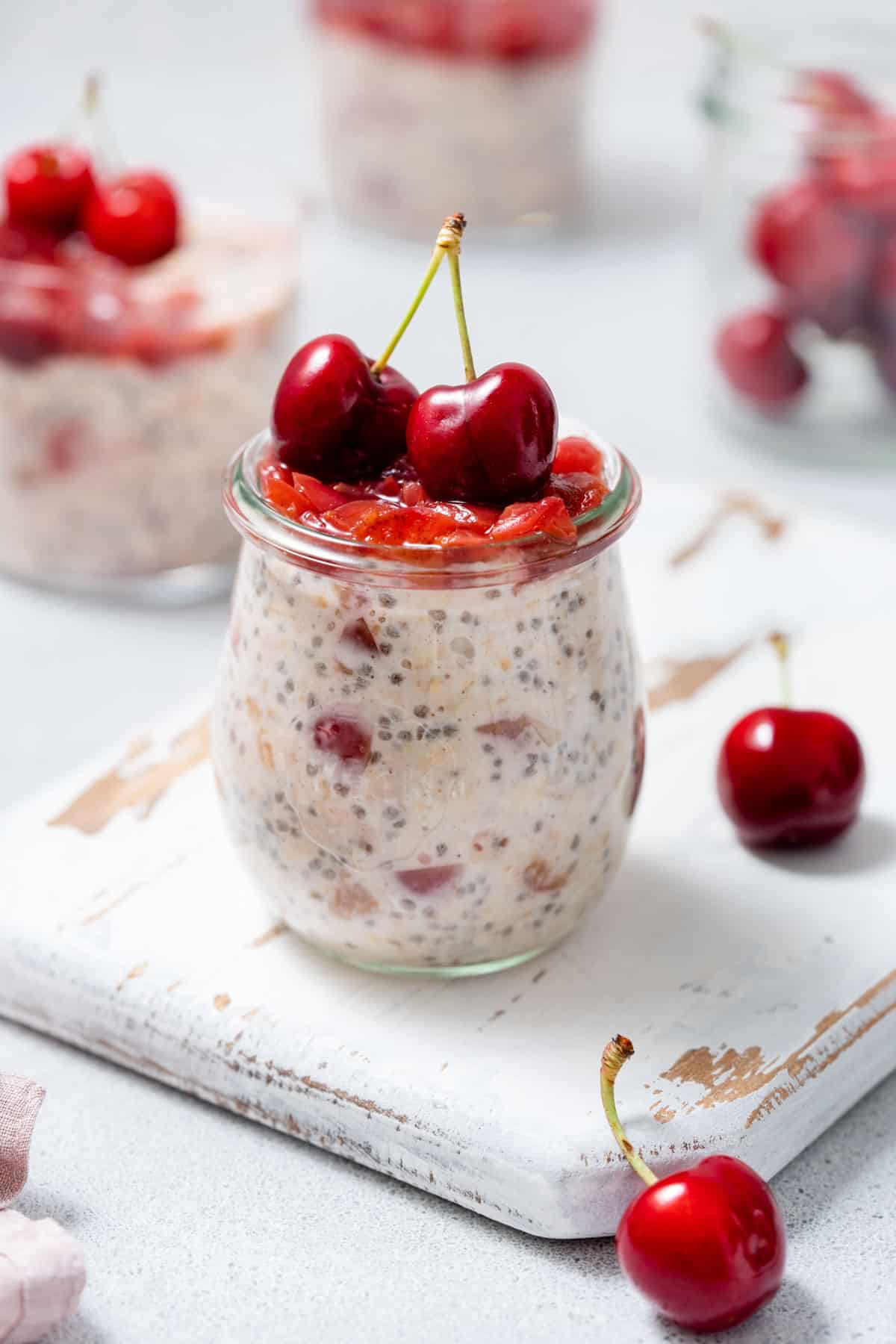 A jar of cherry overnight oats with cherry jam and fresh cherries on top on a serving board with fresh cherries scattered.