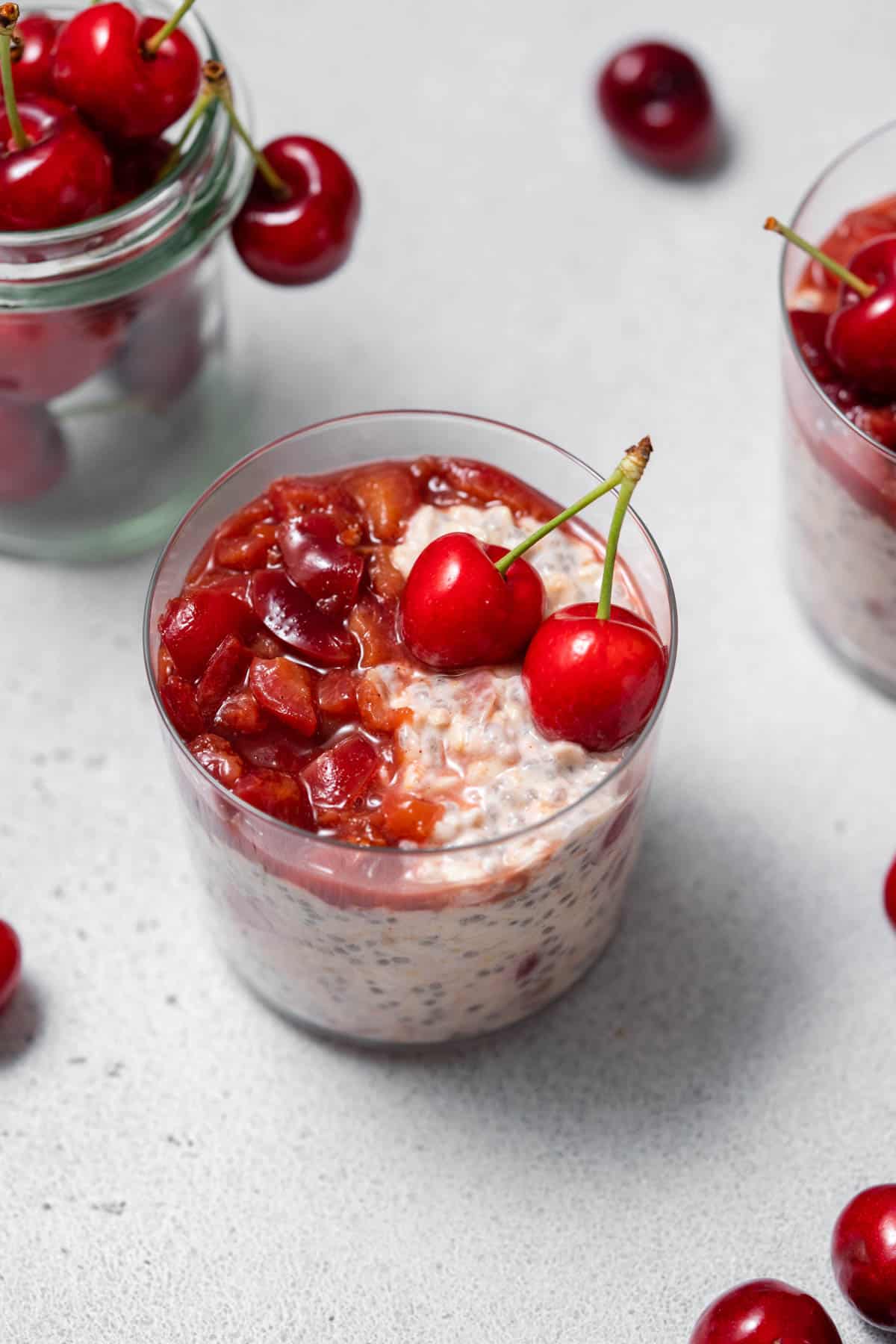 A slightly overhead view of a glass of cherry overnight oats with cherry jam and fresh cherries on top.
