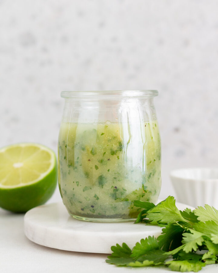 A jar of cilantro lime vinaigrette on a coaster with some cilantro and a cut lime around it.