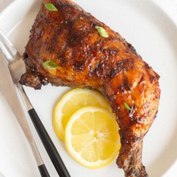 A grilled chicken leg quarter on a plate with two slices of lemon and a fork and knife beside it.