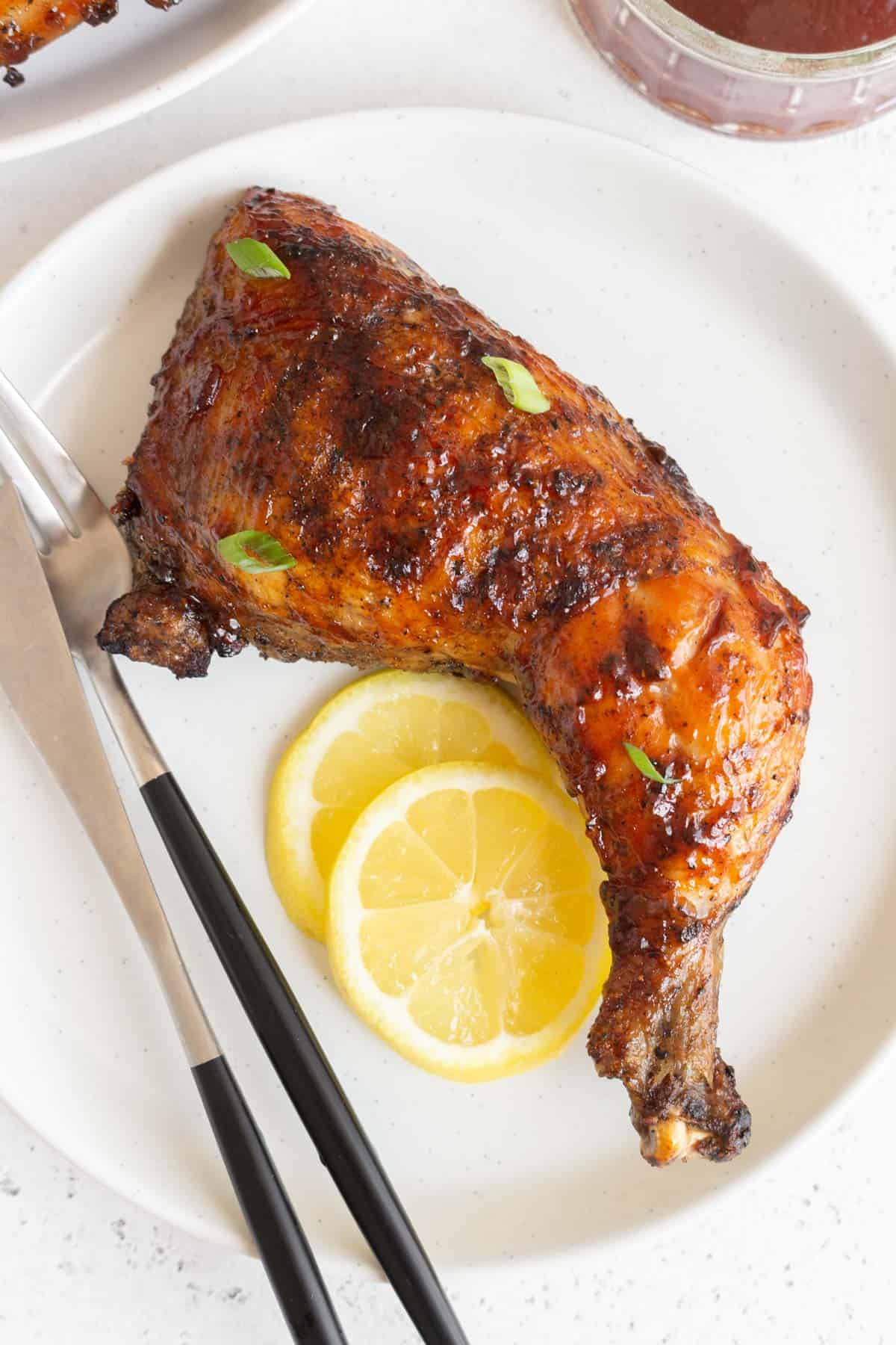 A grilled chicken leg quarter on a plate with two slices of lemon and a fork and knife beside it.