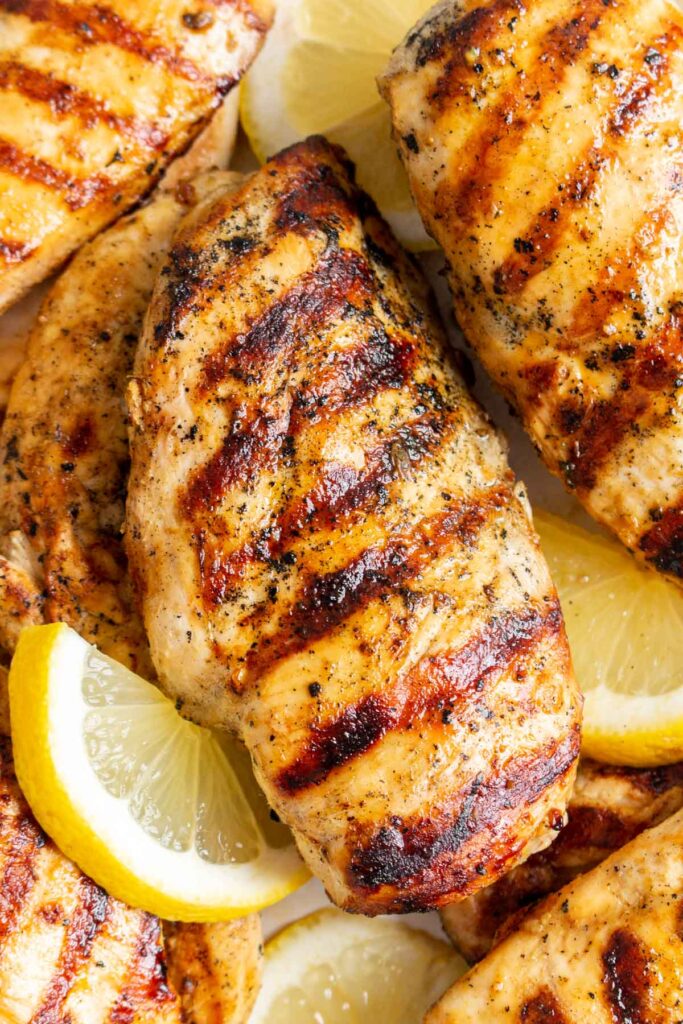 Close up of a grilled lemon pepper chicken breast with grill marks surrounded by more chicken and lemon slices.