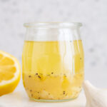 A jar of lemon garlic vinaigrette on a coaster with a lemon and garlic around it.