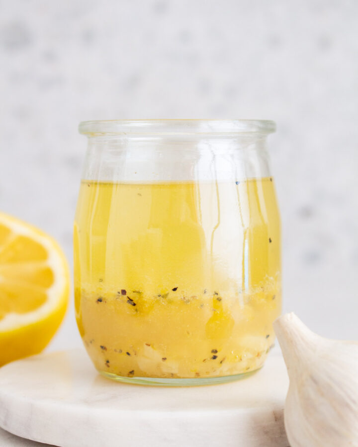 A jar of lemon garlic vinaigrette on a coaster with a lemon and garlic around it.