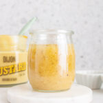 A jar of maple dijon dressing on a coaster with a jar of dijon in the background.