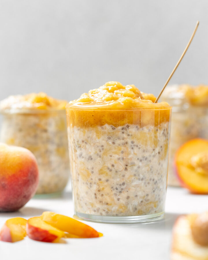 A glass with peach overnight oats topped with cooked peaches with cut peaches around it.