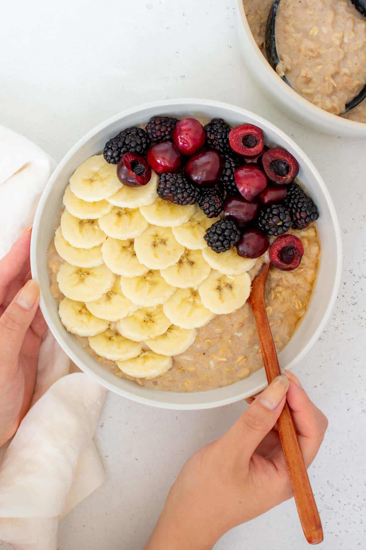 A bowl of egg white oatmeal topped with fresh berries and banana slices with a hand holding a spoon tucked into it.