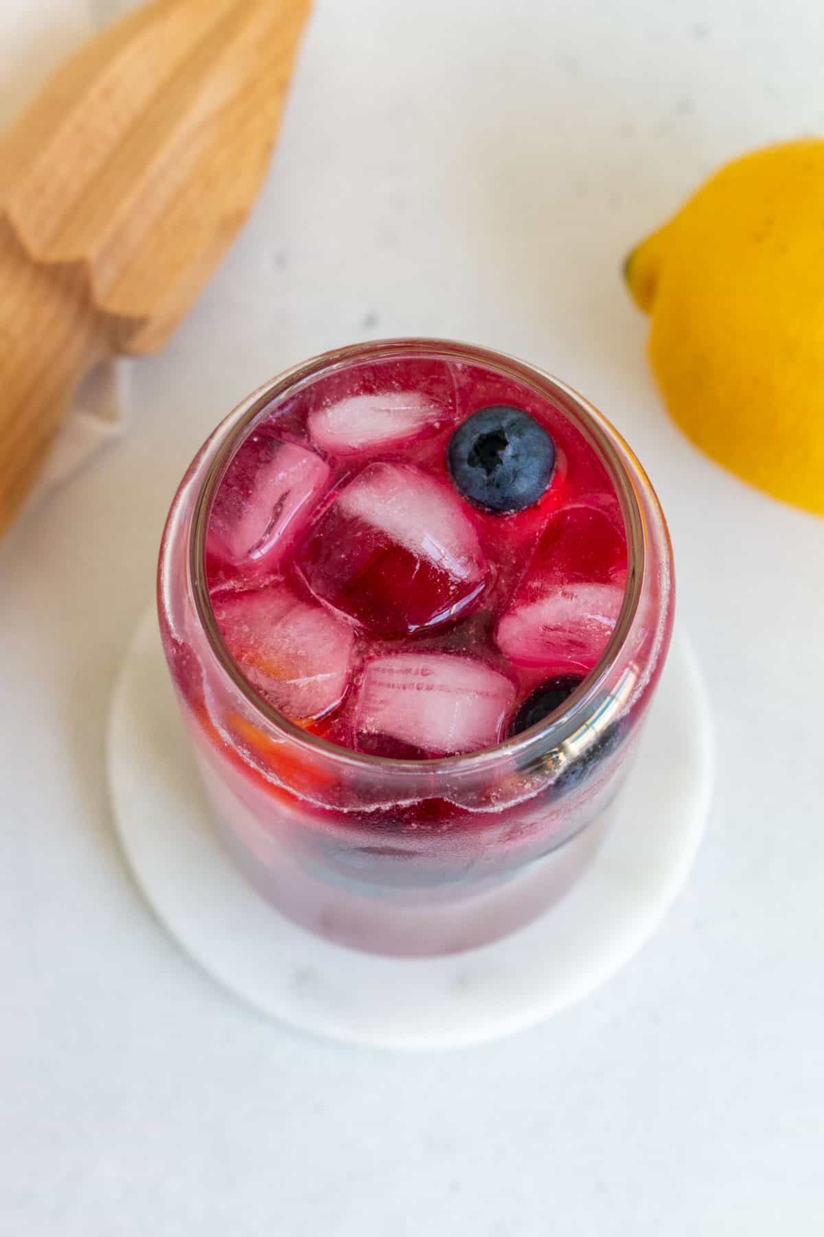 An angled view of a glass of blueberry lemonade with ice and fresh blueberries and lemon slices.