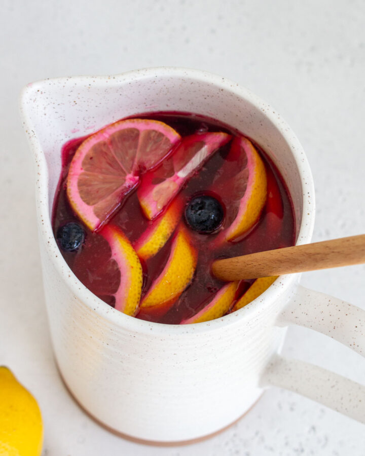 An angled view of a pitcher of blueberry lemonade with a wooden spoon inside.
